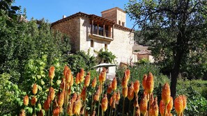 Um edifício de pedra com varanda, rodeado por vegetação exuberante e plantas floridas.