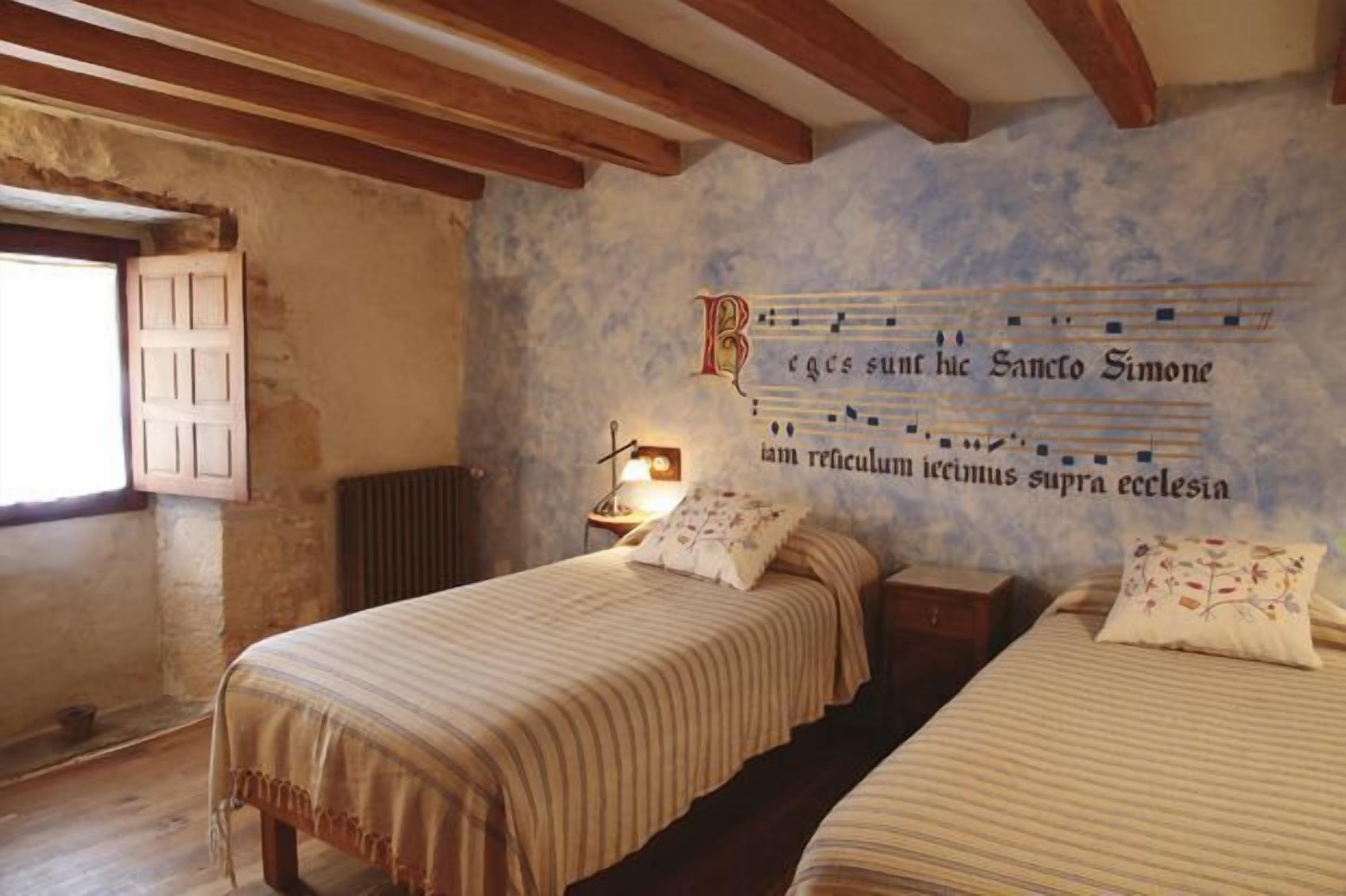 A bedroom with two beds, wooden beams on the ceiling, a wall with a religious inscription, and a window with a wooden shutter.