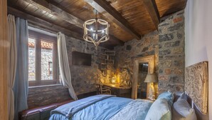 A rustic bedroom with a stone wall, wooden beams, a bed with blue bedding, a desk with a lamp, and a window with curtains. - Dandy Villas Palios Agios Athanasios | Attic | Fireplace | Up to 8 (Agios Athanasios)