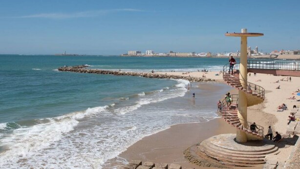 A beach with a spiral staircase leading to a viewing platform, a sandy shore, and a distant cityscape. - Apartamento El rosal de Huertas (Cádiz)