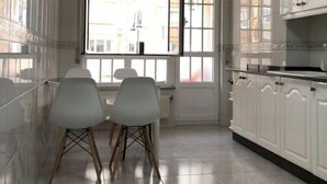 Een keuken met witte kastjes, een eettafel met stoelen en een raam met uitzicht op gebouwen.