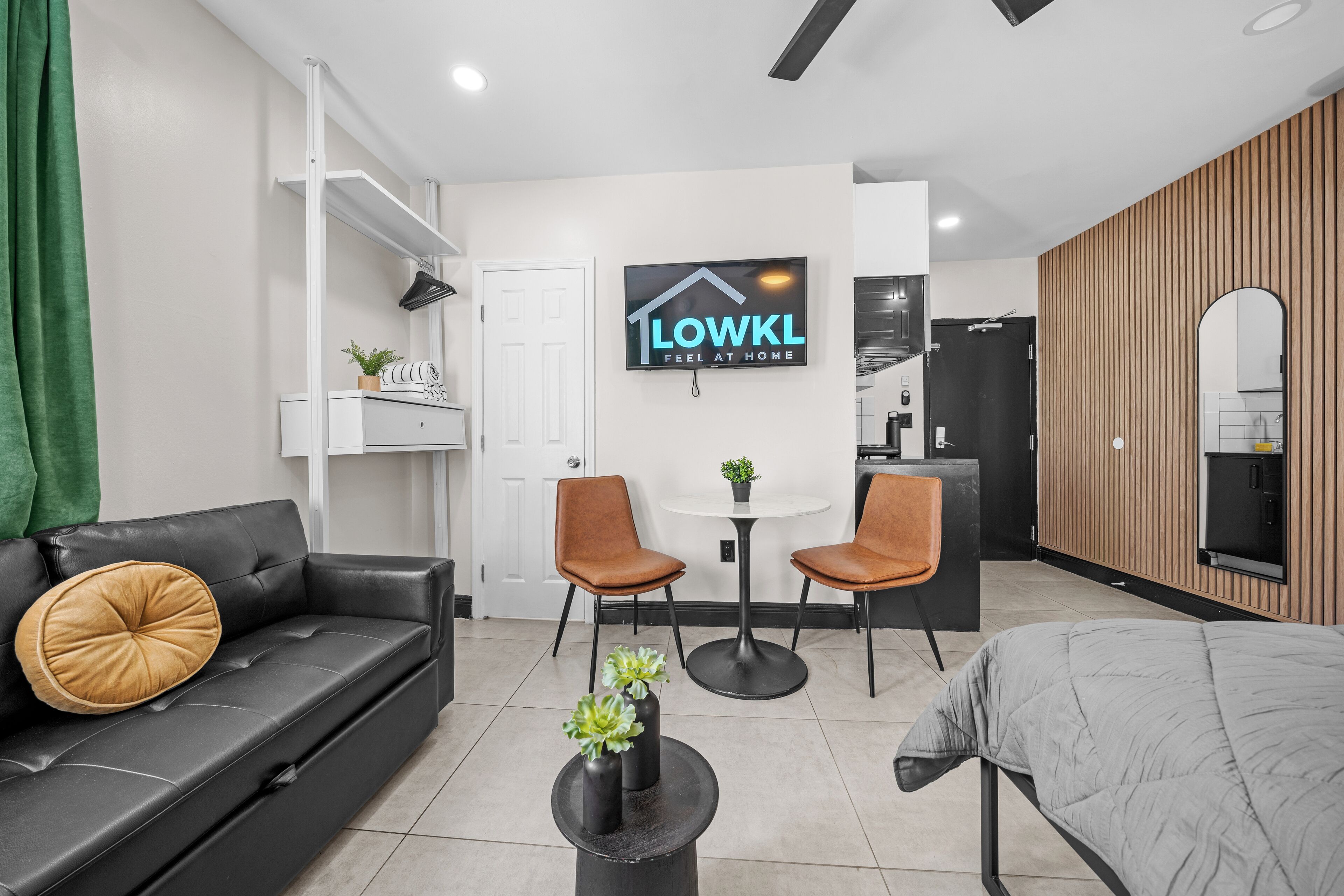A modern living room with a black leather sofa, two brown chairs, a small round table, and a TV mounted on the wall.