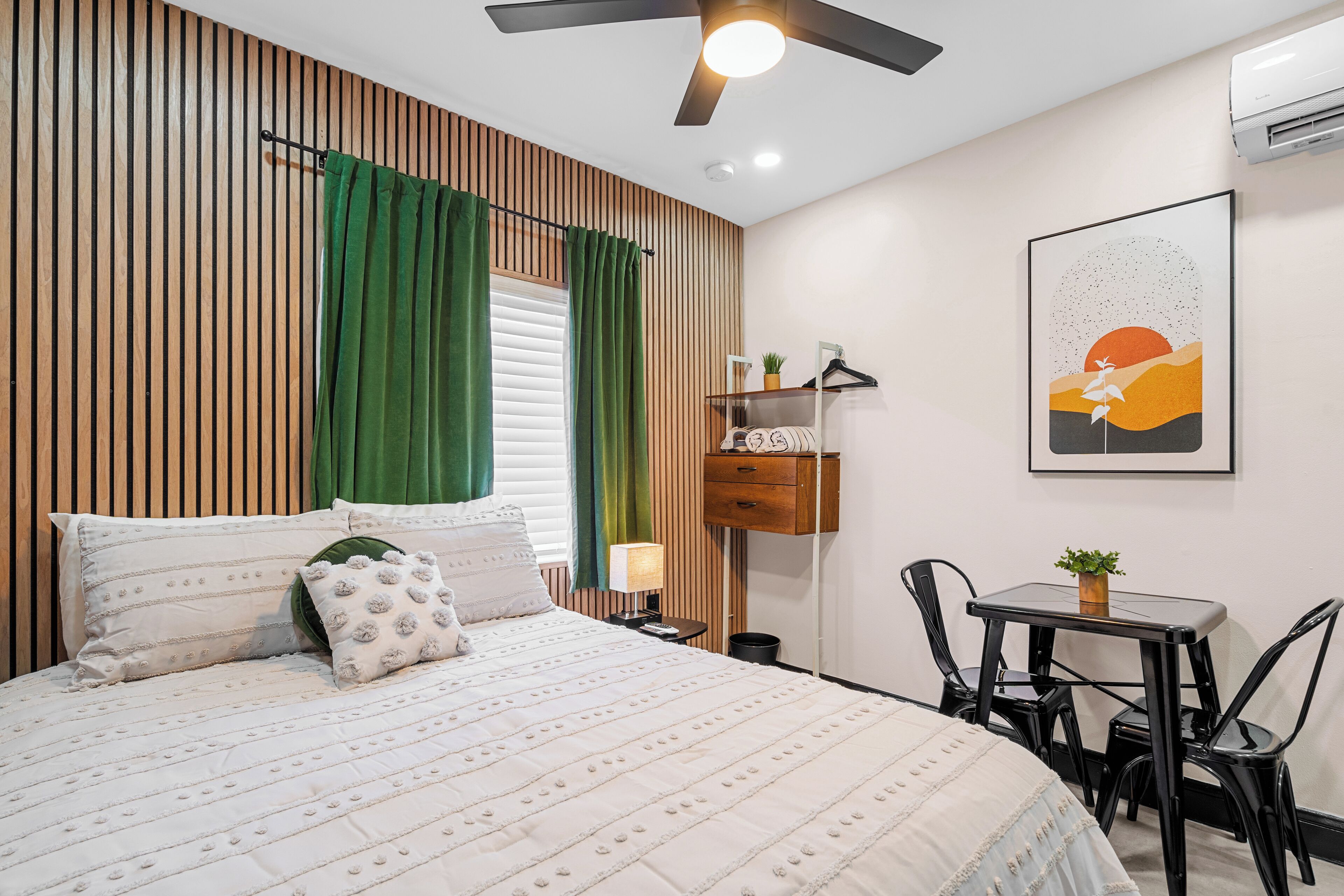 A bedroom with a bed, a desk, chairs, a wall-mounted shelf, and a ceiling fan.