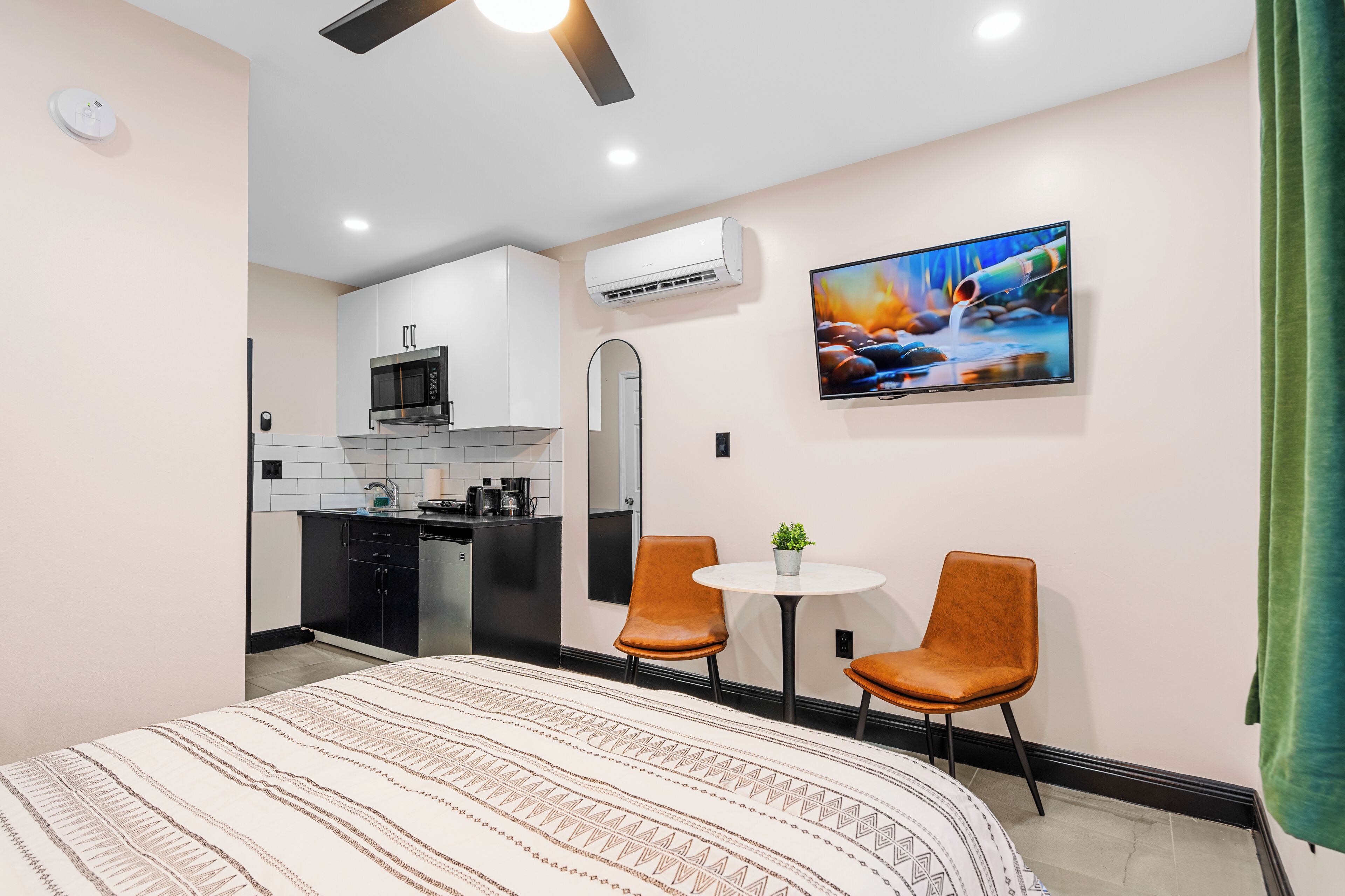 A modern hotel room with a bed, a small kitchenette, two chairs, a TV, and a painting on the wall.