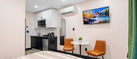 Una habitación de hotel moderna con una cama, una kitchenette pequeña, dos sillas, un televisor y un cuadro en la pared.