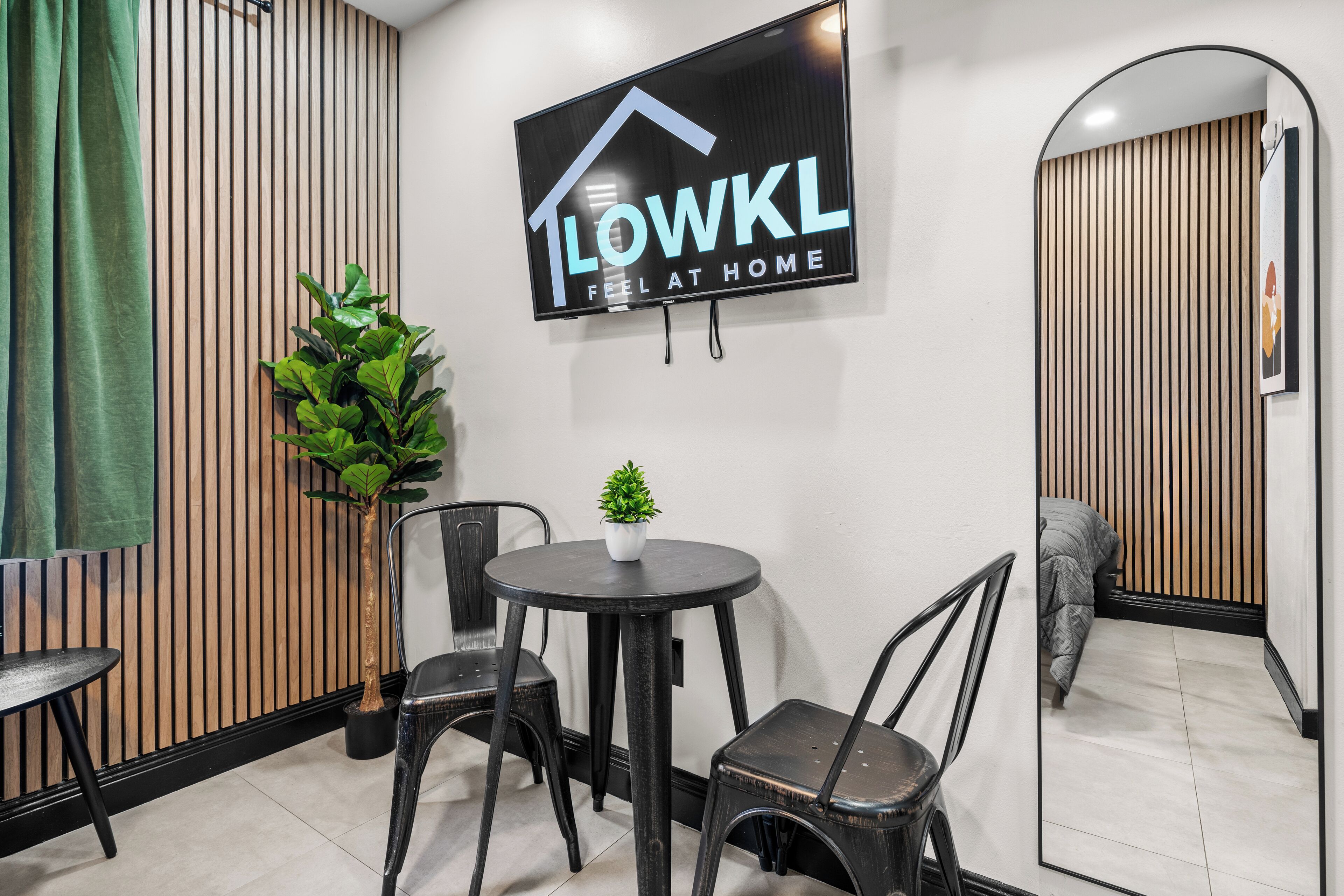 A modern interior with a mounted TV displaying "LOWKL FEEL AT HOME", a round table with chairs, a potted plant, and a mirror reflecting a bed and a wooden wall.