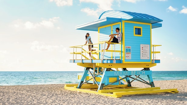 A lifeguard tower with a blue and yellow color scheme, labeled "MIAMI BEACH," on a sandy beach with the ocean in the background. - Baybreeze Apartments by Lowkl (Miami Beach)