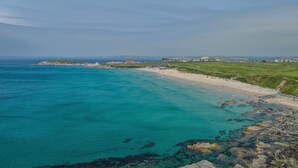 Un paysage côtier avec une plage, des eaux turquoise et limpides, et un ensemble de bâtiments situé au loin.