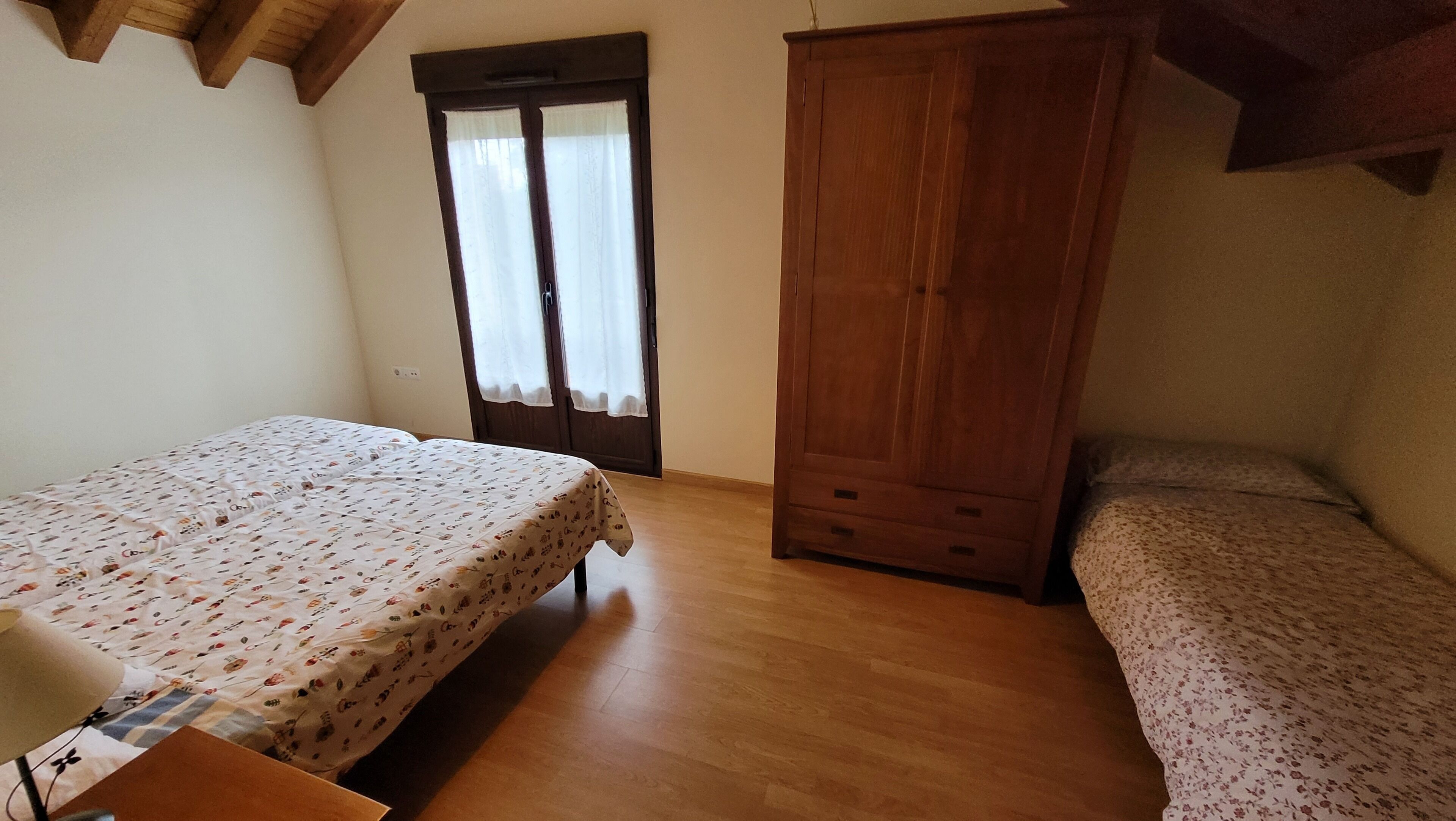 A bedroom with two beds, wooden wardrobe, and a window with curtains.