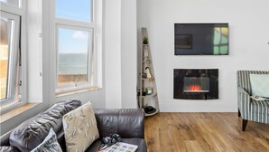 Un salon avec un canapé en cuir, une cheminée, un téléviseur fixé au mur et une vue sur la mer grâce à de grandes fenêtres.