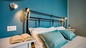 Un lit bien fait, avec deux oreillers, une table de chevet sur laquelle repose un livre ouvert et un luminaire fixé au mur.