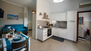 Une cuisine compacte avec des meubles blancs, un four à micro-ondes et une cuisinière. On y trouve un coin repas avec une table dressée pour deux personnes, un canapé bleu et un tableau accroché au mur.