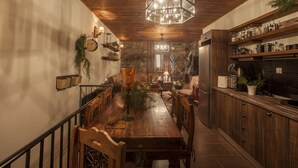A cozy dining area with a wooden table, chairs, and a chandelier. There are shelves with kitchen items and a refrigerator in the background. - Dandy Villas Palios Agios Athanasios |Rustic | Fireplace | Up to 6 (Agios Athanasios)