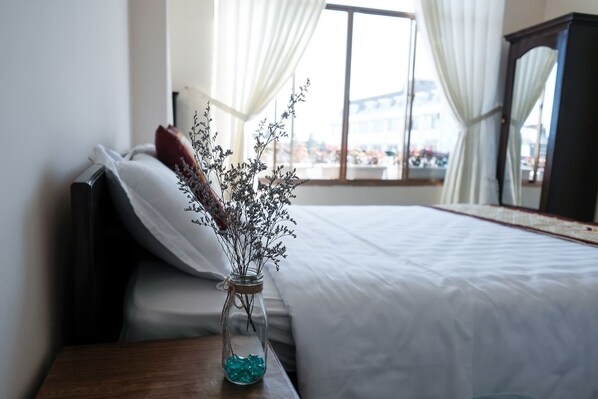 A bedroom with a bed, a nightstand, a vase with dried flowers, and a window with curtains.
