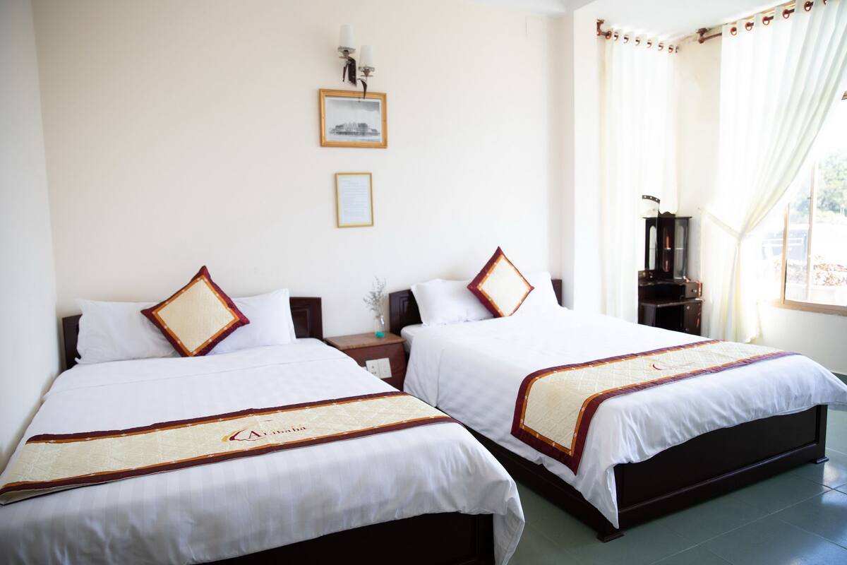 A hotel room with two beds, each with a white bedspread and a decorative border. There is a framed picture on the wall and a nightstand with a lamp between the beds.