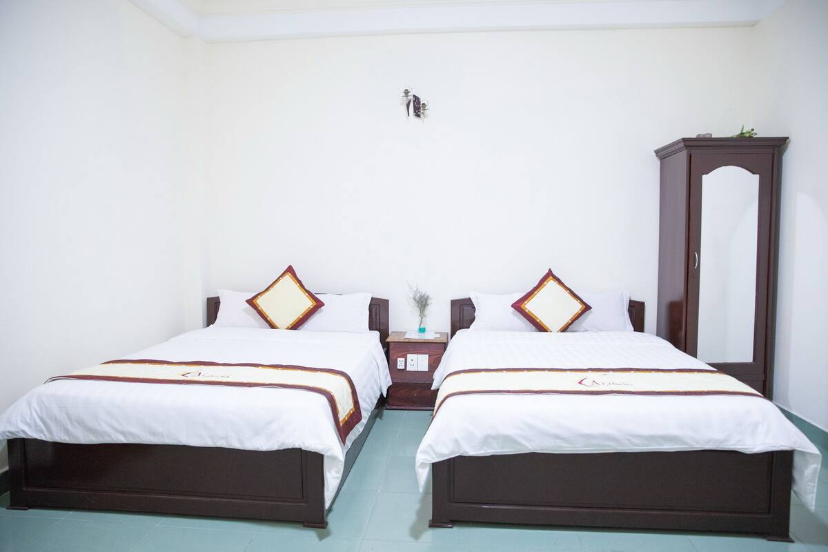 A hotel room with two single beds, a wooden wardrobe, a nightstand with a vase, and a wall-mounted light fixture.