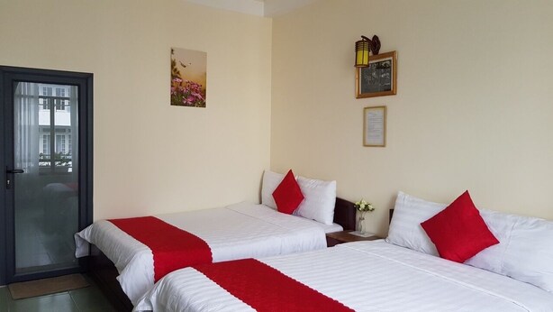 Une chambre d’hôtel avec deux lits, chacun agrémenté de draps blancs et d’oreillers rouges, une petite table sur laquelle est posé un vase de fleurs, et une porte dotée d’un panneau de verre.