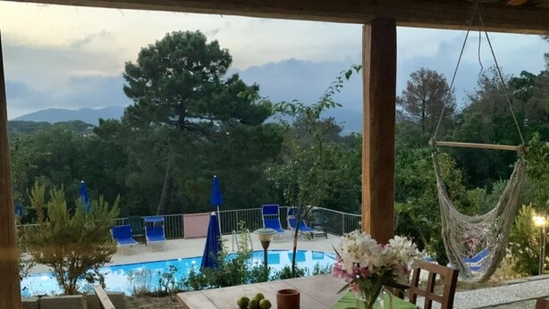 An outdoor dining area with a wooden table, chairs, and a hanging hammock. In the background, there is a pool surrounded by blue lounge chairs and a view of trees and mountains. - Apt. Sera & Mattino - SERAMATTINO B (Capoliveri)