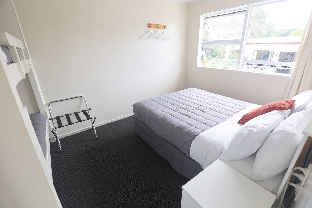 A small, neatly arranged bedroom with a bed, a small table, a chair, and a window providing a view of the outside.