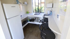 A compact kitchen with a refrigerator, sink, and stove. There are two black chairs and a window with a view of greenery outside. - Orere Point TOP 10 Holiday Park (Orere Point)