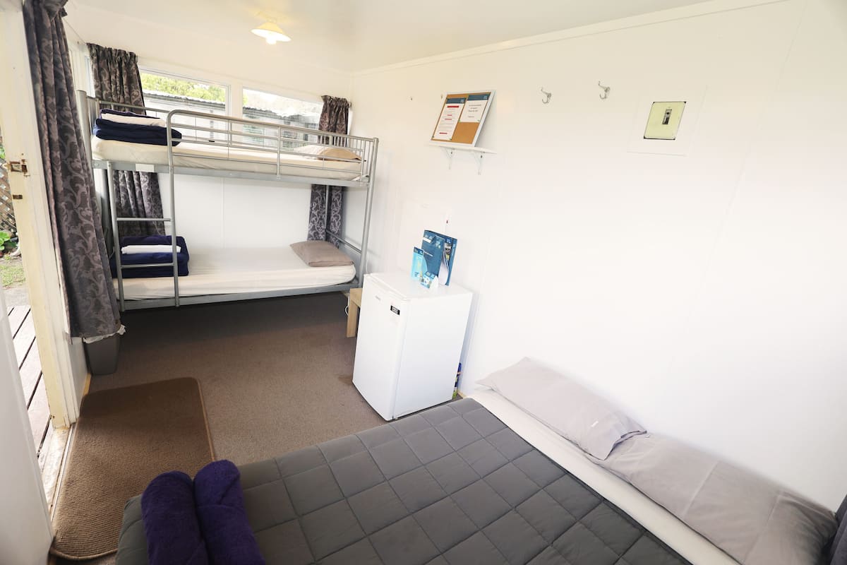 A bunk bed room with a small white fridge, a bedside table, and a window with curtains.