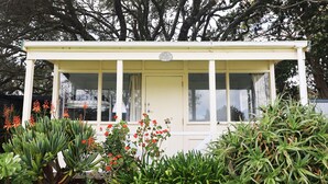 A small, single-story building with a white exterior and a door. It is surrounded by various plants and flowers, including red flowers and green foliage. - Orere Point TOP 10 Holiday Park (Orere Point)