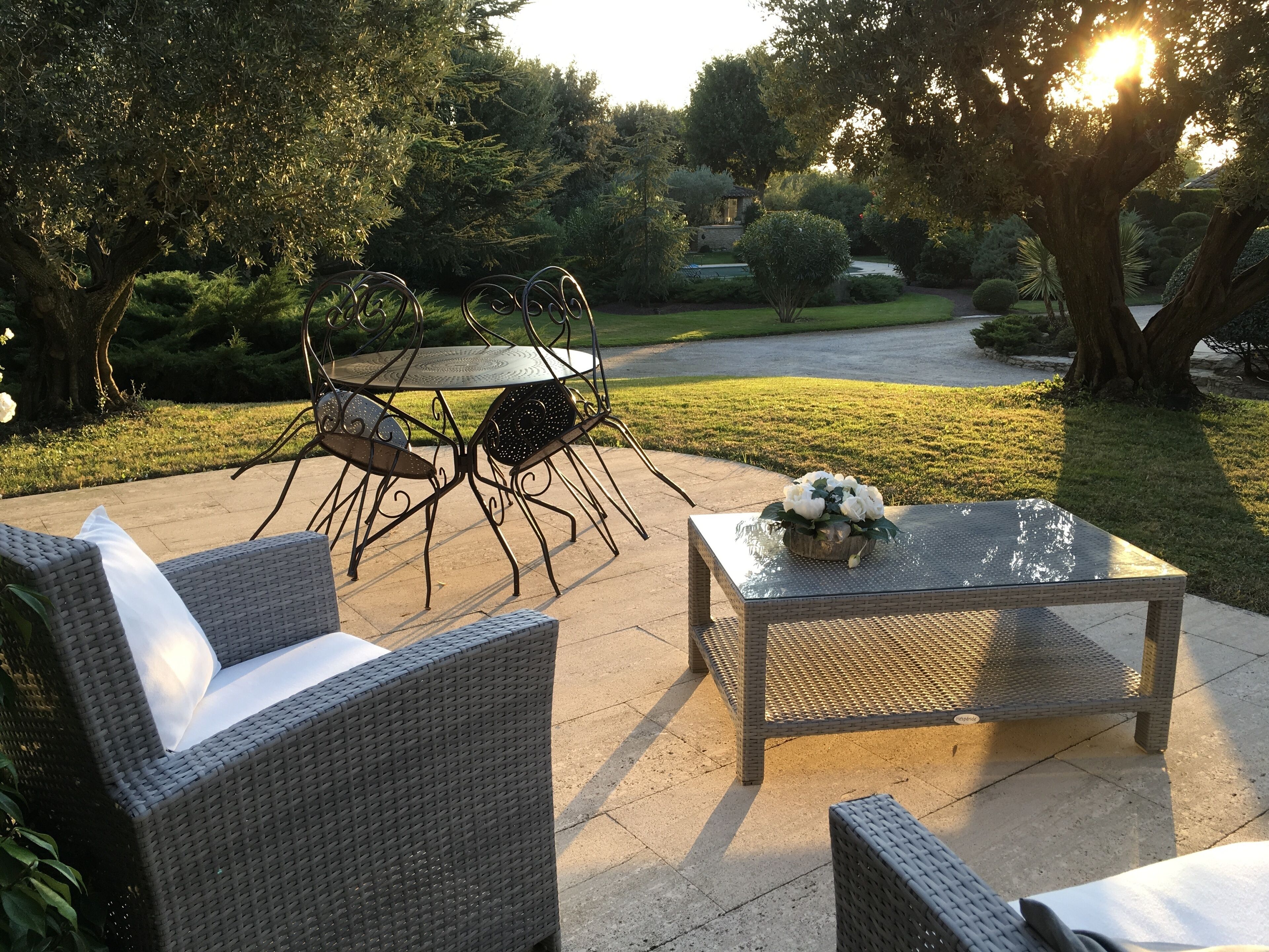 Un patio esterno con mobili in vimini, un tavolo in vetro e un centrotavola floreale, con vista su un giardino curato.