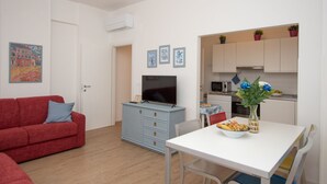 A compact living space with a red sofa, a TV on a blue cabinet, a dining table with a bowl of fruit, and a kitchen area with cabinets and a microwave. - 65 sqm apartment 50 meters from the sea (Albissola Marina)