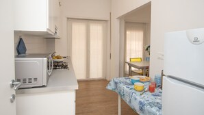 A compact kitchen with a microwave, gas stove, and a refrigerator. There is a dining area with a table and chairs, and a window with curtains. - 65 sqm apartment 50 meters from the sea (Albissola Marina)