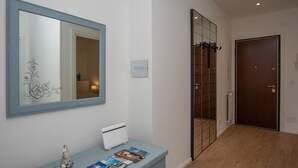 A hallway with a wooden floor, a blue-framed mirror, a white console table with magazines, and a brown door. - 65 sqm apartment 50 meters from the sea (Albissola Marina)