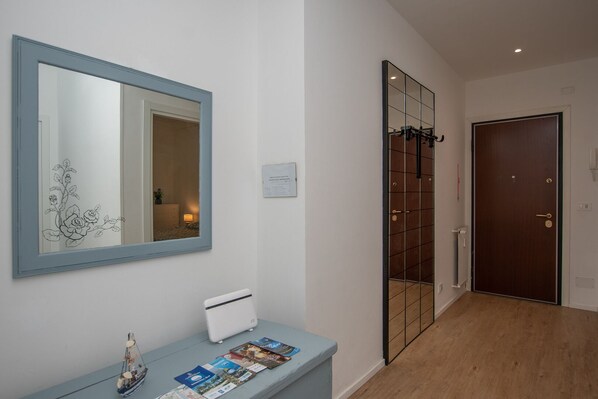 A hallway with a wooden floor, a blue-framed mirror, a white console table with magazines, and a brown door.