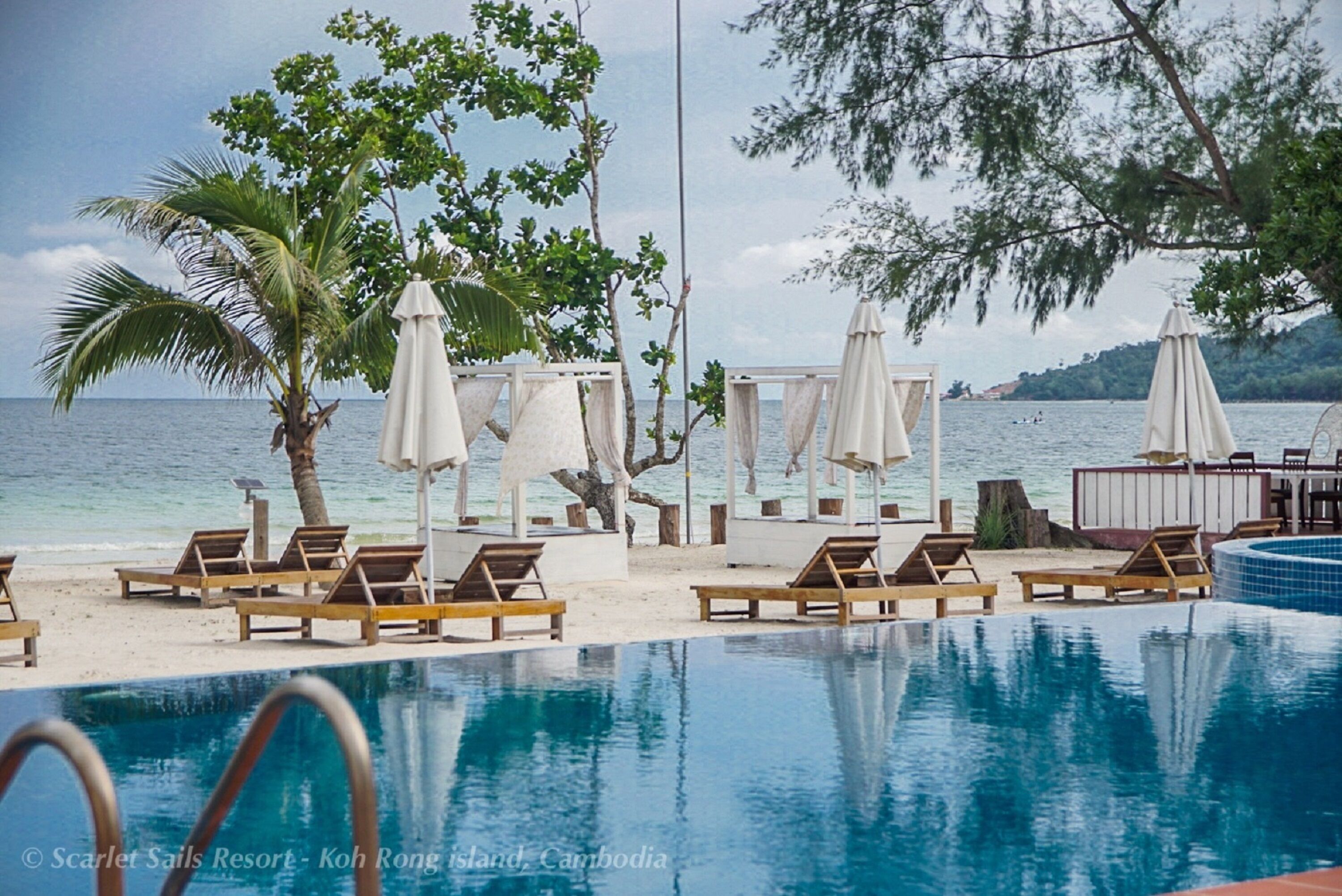 Zona junto a la piscina con sillones de descanso y sombrillas, vista despejada al océano y entorno tropical.