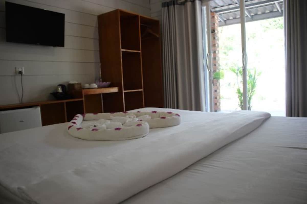 A hotel room with a bed, a wooden wardrobe, a flat-screen TV mounted on the wall, and a window with curtains.