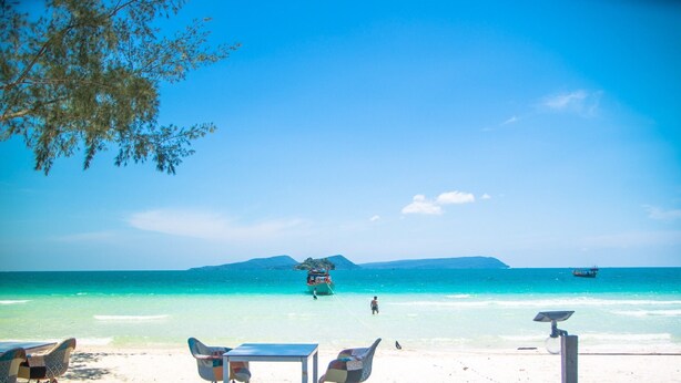 A beach view with clear turquoise water, a boat in the distance, and a person walking on the shore. - Scarlet Sails Resort (Koh Rong)