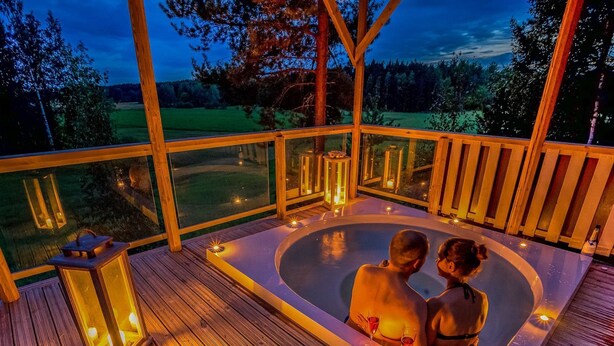 A couple in a hot tub with a wooden deck and glass walls, overlooking a grassy landscape. - Villa ebba by Interhome (Sysmä)
