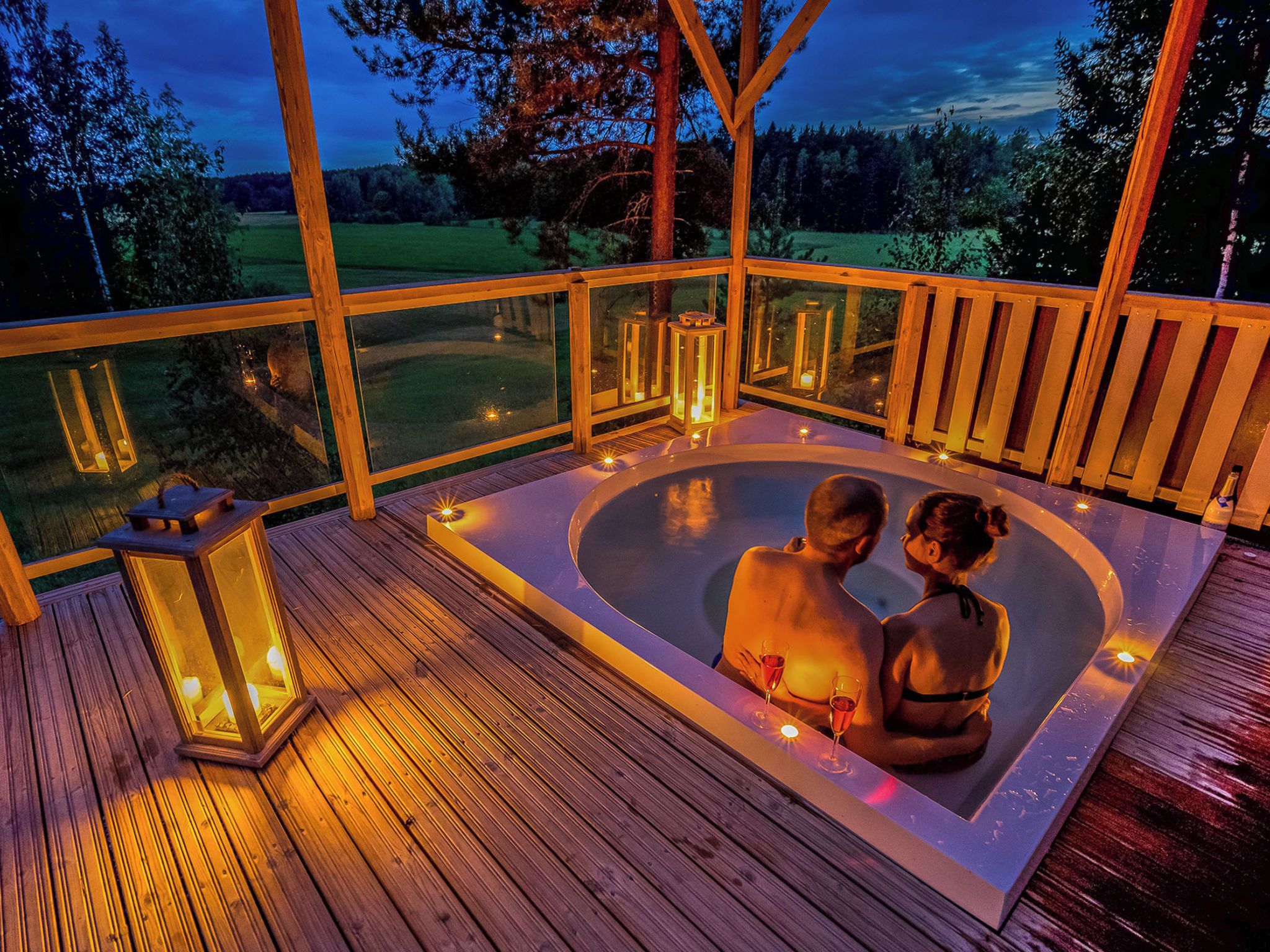 Una pareja en un jacuzzi con una terraza de madera y barandas de vidrio, con vistas a un paisaje cubierto de césped.