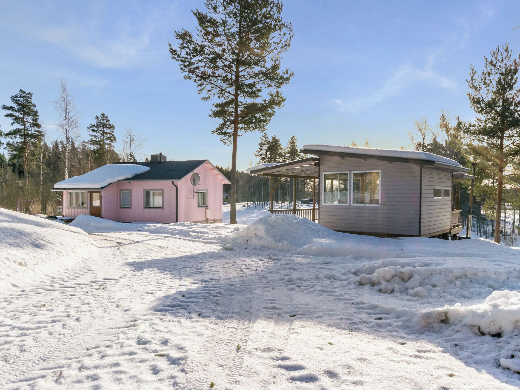 Schneebedeckte Landschaft mit zwei Hütten, eine rosa und eine grau, umgeben von Bäumen.