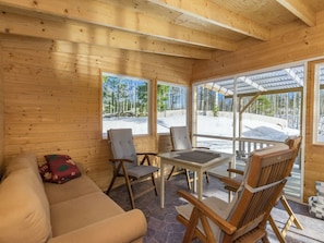 Interior de uma cabana de madeira com sofá, cadeiras, mesa e janelas amplas que exibem uma paisagem nevada.