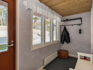 A room with a wooden ceiling, a window with a white valance, a radiator, a round stool, and a coat rack with a jacket and an umbrella.