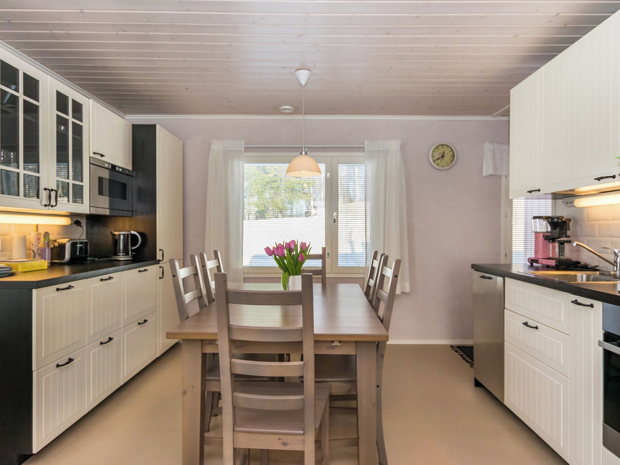 Een kleine keuken met witte kastjes, een houten eettafel en een raam met witte gordijnen.