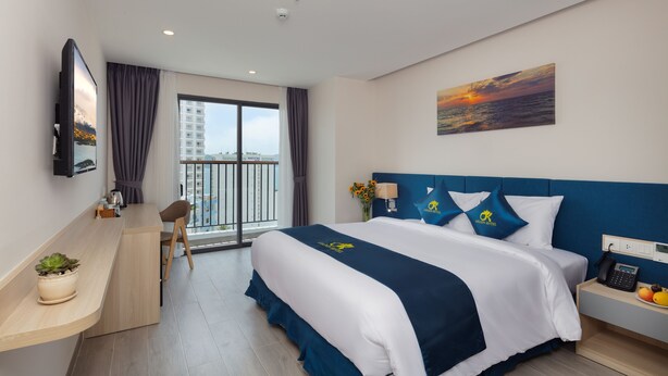 A modern hotel room with a large bed, a desk, a chair, a TV, a painting, and a balcony with a city view. - CTK Golden Hotel (Nha Trang)