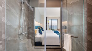 A modern hotel bathroom with a glass shower enclosure, a visible bedroom with a bed and a view of the sea, and a towel rack with a white towel. - CTK Golden Hotel (Nha Trang)
