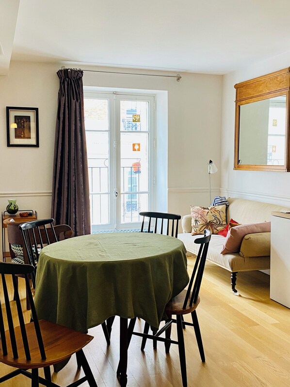 Uma sala de jantar com uma mesa redonda coberta por toalha verde, rodeada por cadeiras de madeira. Há uma janela com cortinas, um quadro na parede e uma luminária sobre uma mesinha lateral.
