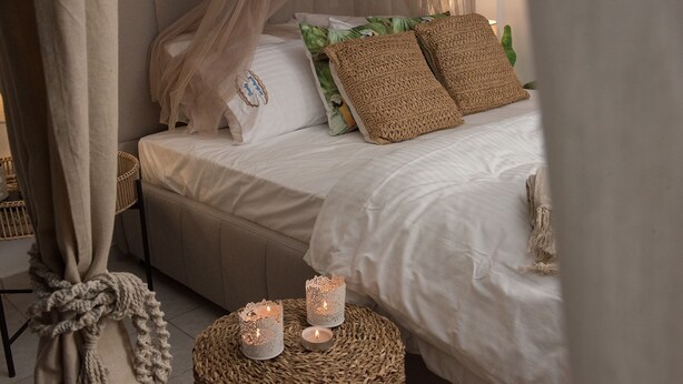 Camera da letto con un letto, biancheria bianca e due candele decorative su uno sgabello in vimini.