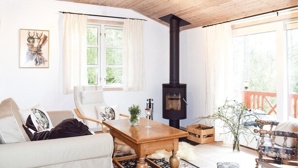 Una habitación acogedora con techo de madera, un ventanal con cortinas, un sofá blanco con cojines decorativos, una mesa de madera con un jarrón y una copa, y un estufa de leña de madera negro.