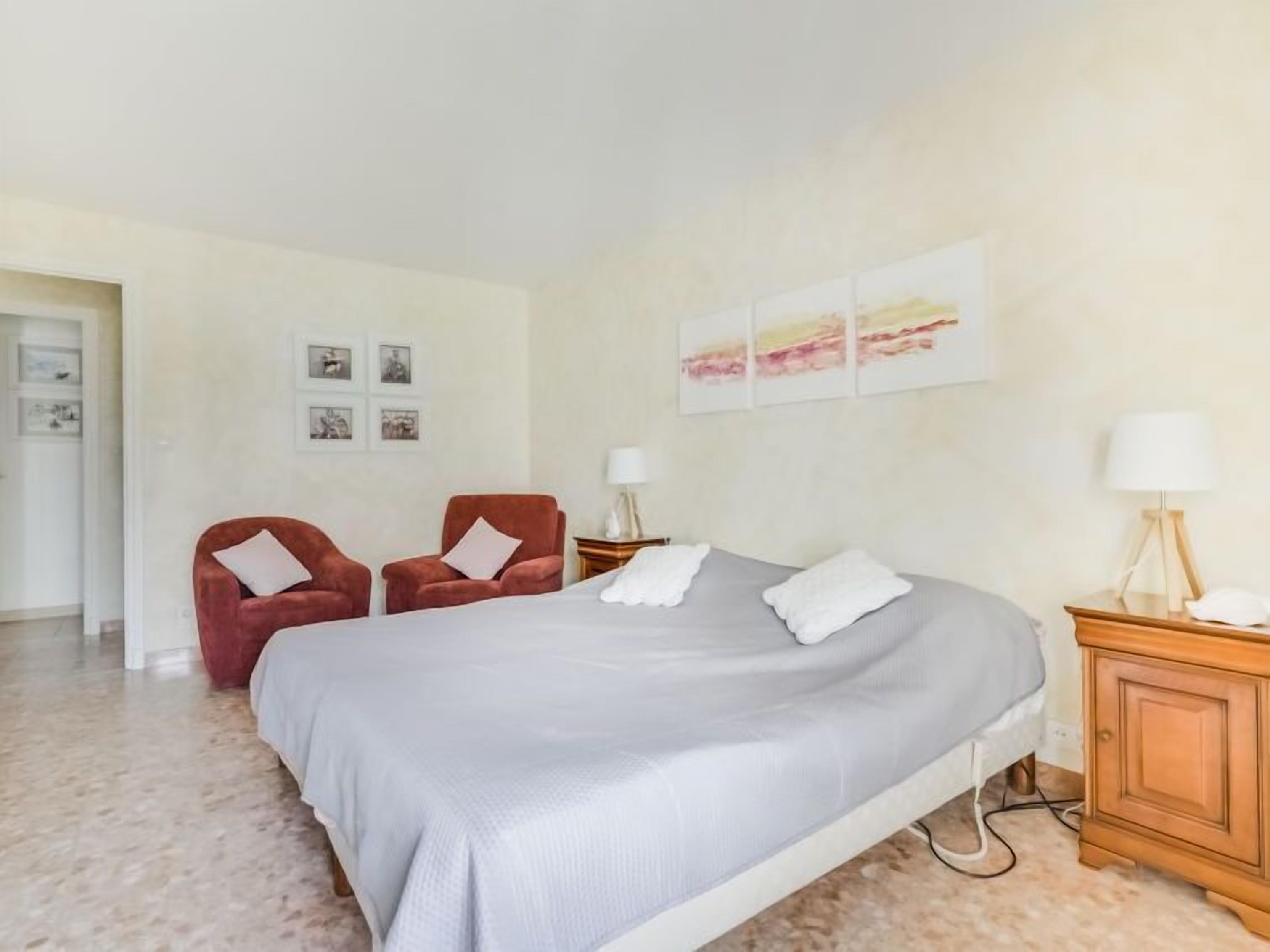 Un dormitorio con una cama, dos sillas rojas, una mesita de noche con una lámpara y una obra de arte en la pared.