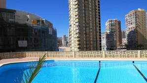 Une piscine dotée d’une terrasse carrelée, entourée d’immeubles de grande hauteur et surplombée par un ciel bleu dégagé.