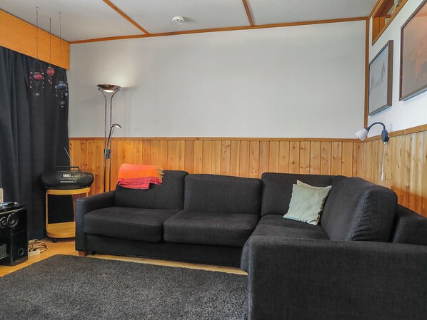 Ein Wohnzimmer mit einem dunklen Sofa, einem kleinen Tisch mit einem Lautsprecher, einer Lampe und Holzvertäfelung.