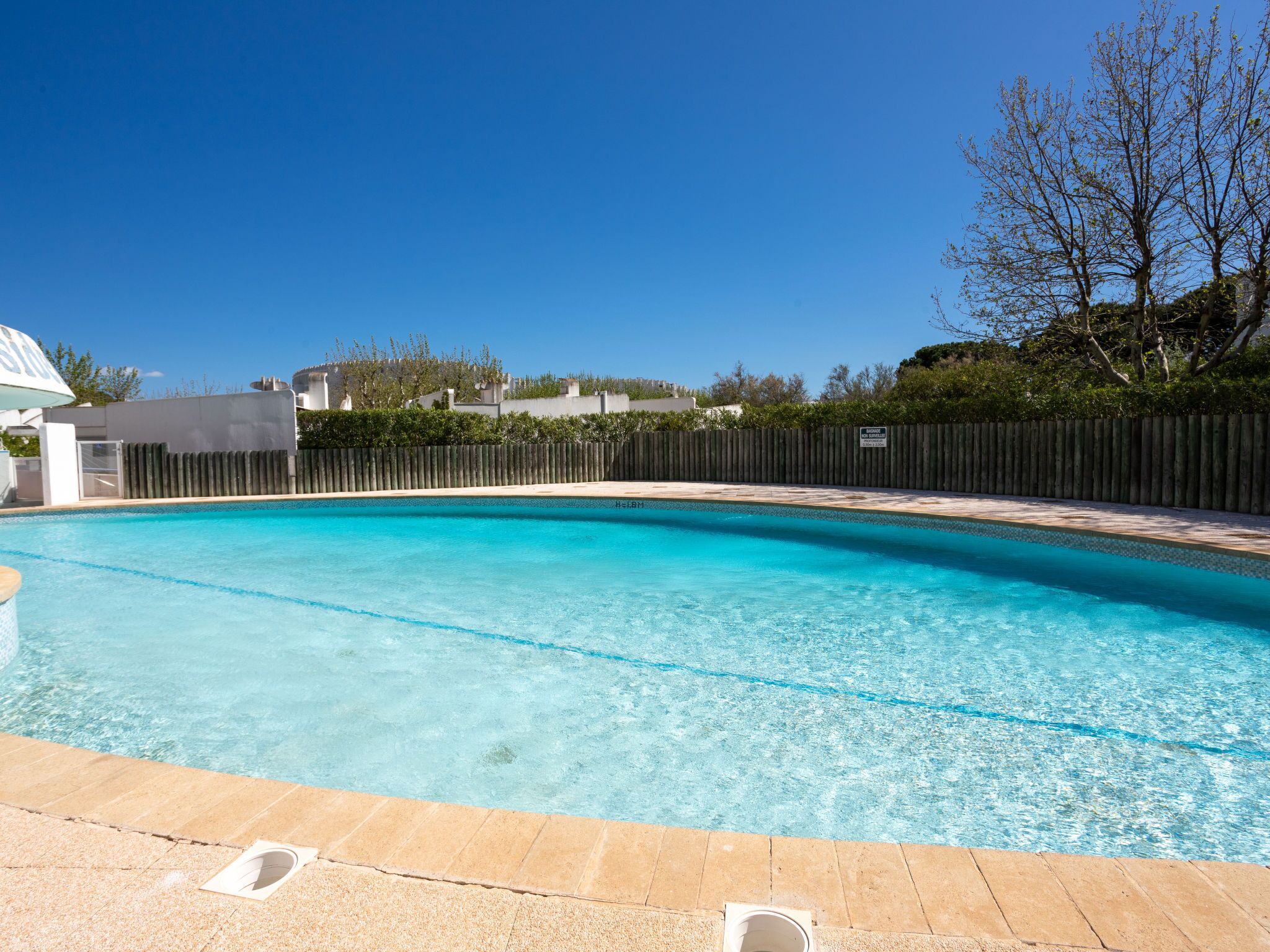 Una piscina rectangular con agua azul cristalina, rodeada por una cerca de madera y un suelo empedrado.