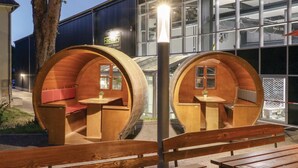 Two wooden barrel-shaped structures with seating and tables, positioned in front of a modern building with large windows. - Amazing home in Thale with sauna (Thale)